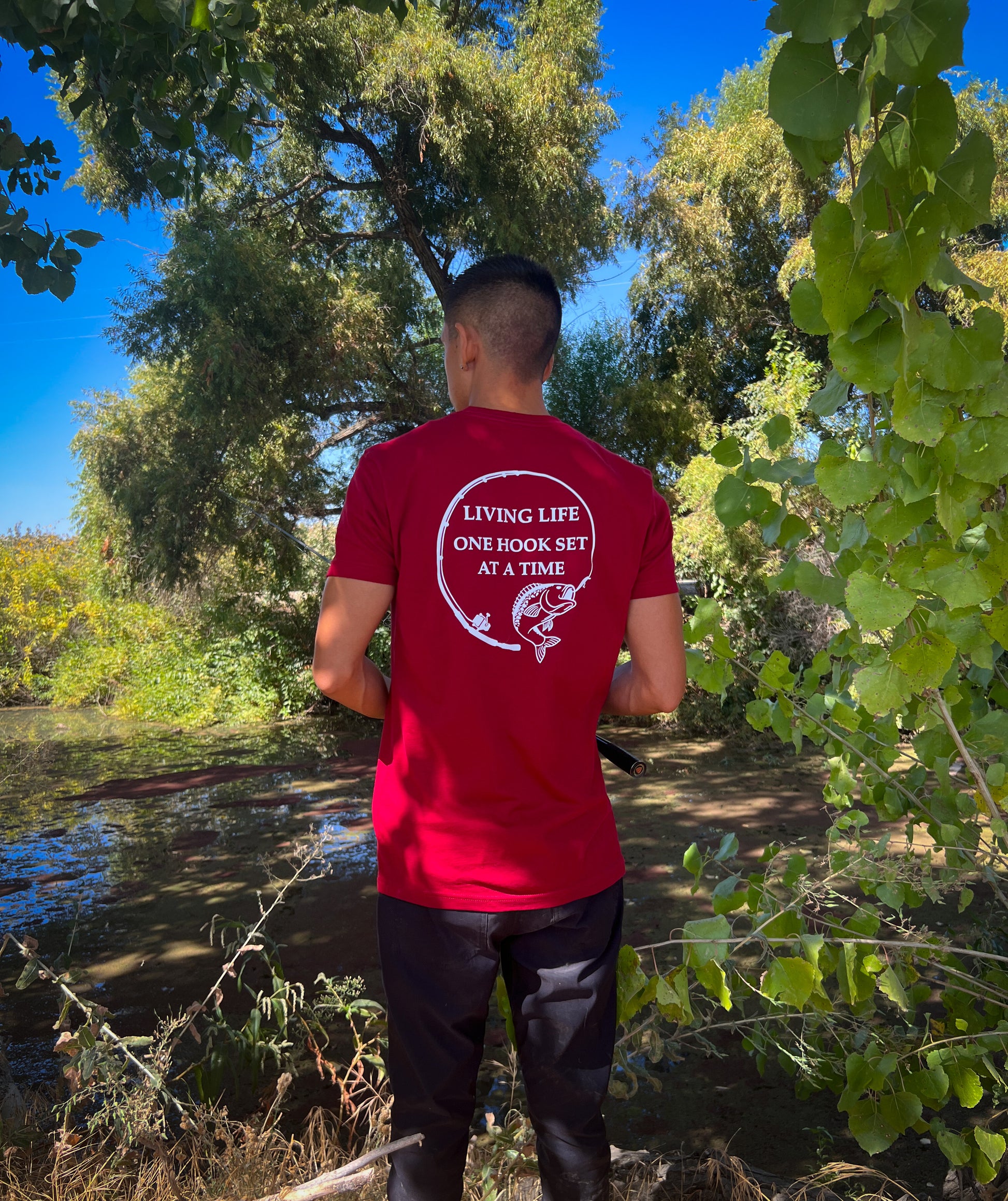 A person standing by a river he has on a burgundy shirt with a design portraying a fishing rod bent in a circle and a large mouth bass at the end of it. In the middle of the design it says Living life one hook set at a time in capital letters