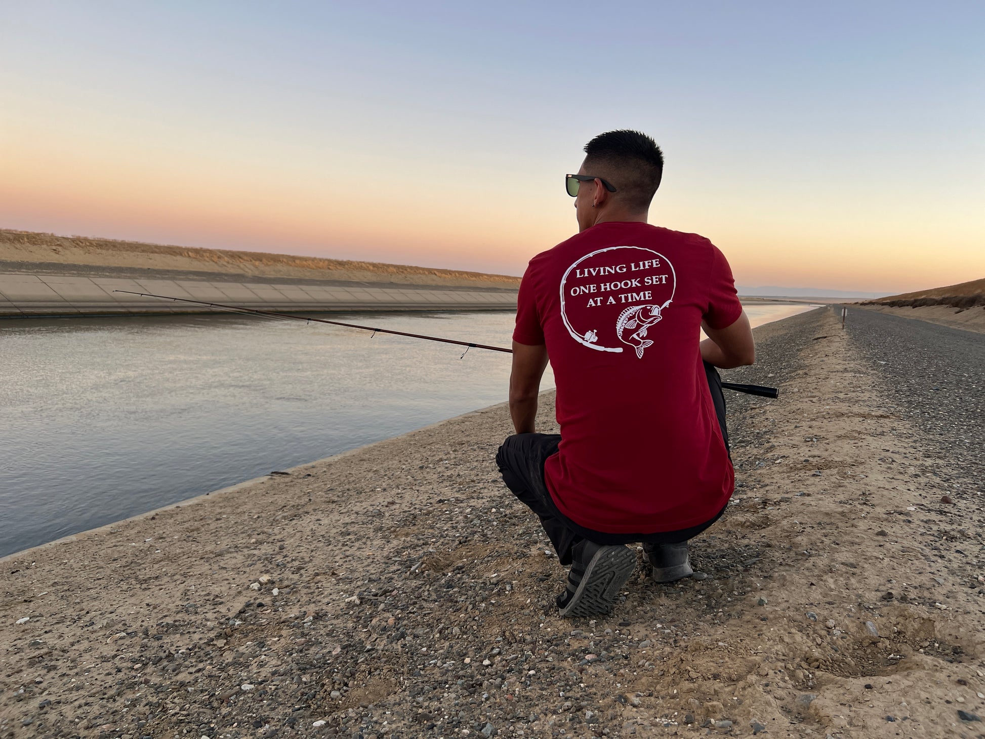 A person kneeling down with a fishing pole. He is next to the California aqueduct and has a burgundy shirt on with a design portraying a fishing rod bent in a circle and a large mouth bass at the end of it. In the middle of the design it says Living life one hook set at a time in capital letters