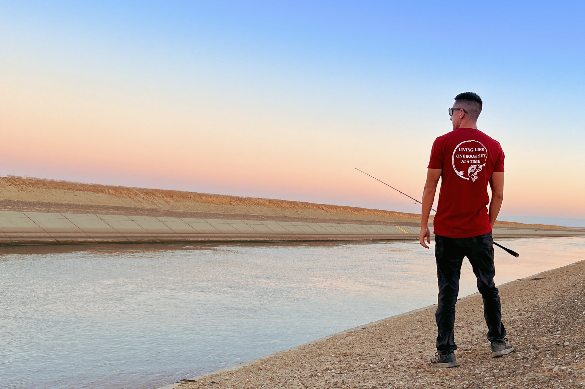 A person standing with a fishing pole. He is next to the California aqueduct and has a burgundy shirt on with a design portraying a fishing rod bent in a circle and a large mouth bass at the end of it. In the middle of the design it says Living life one hook set at a time in capital letters