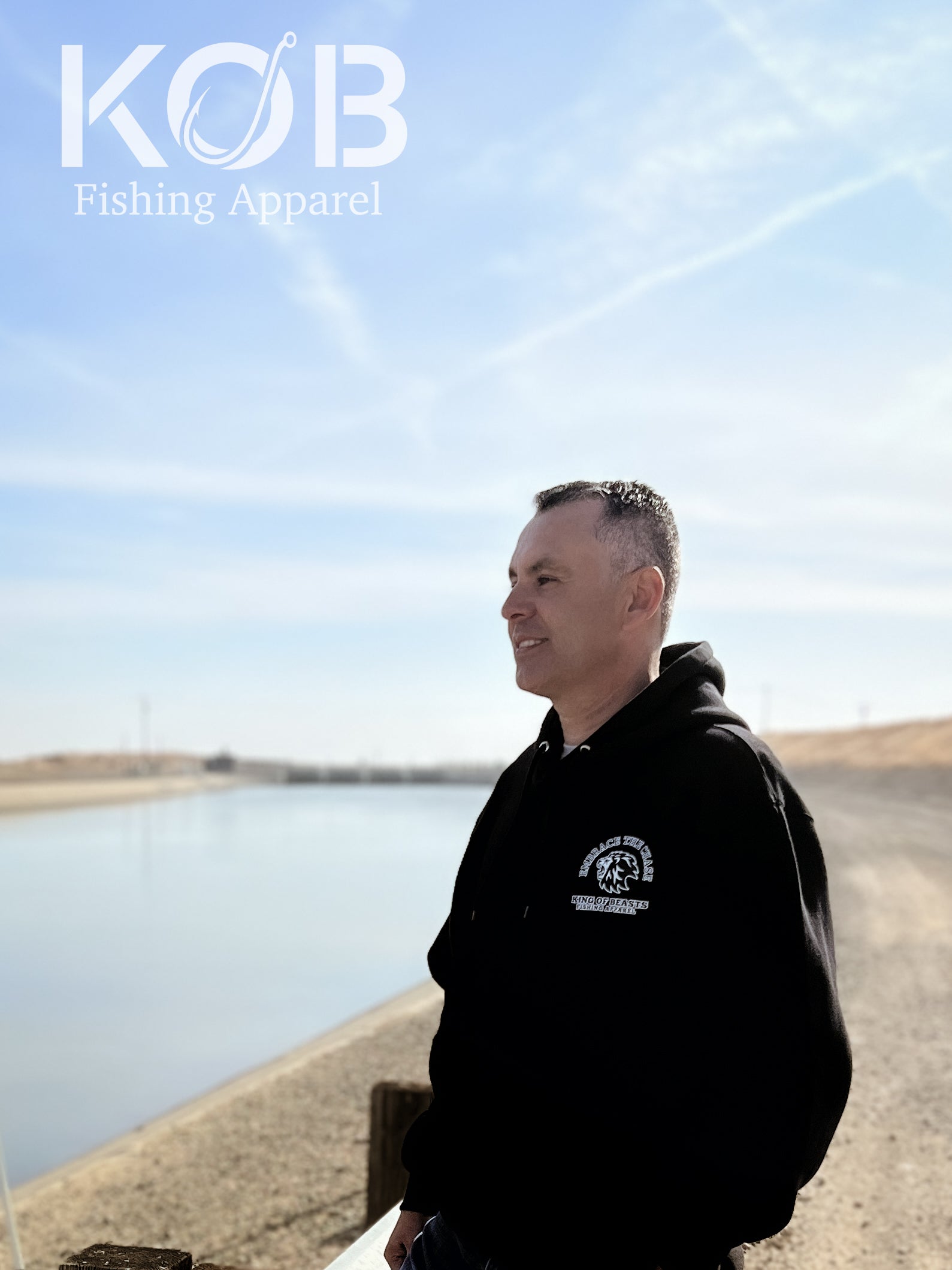 Person standing next to the California aqueduct with a black hoodie on. On the left side of the chest it has a lion roaring and around the lion it says embrace the chase King Of Beasts Fishing Apparel.