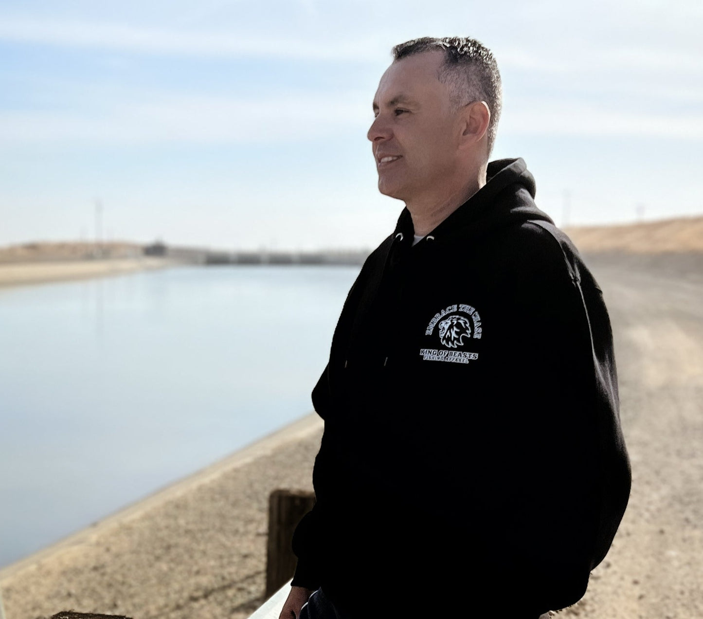 Person standing next to the California aqueduct with a black hoodie on. On the left side of the chest it has a lion roaring and around the lion it says embrace the chase King Of Beasts Fishing Apparel.