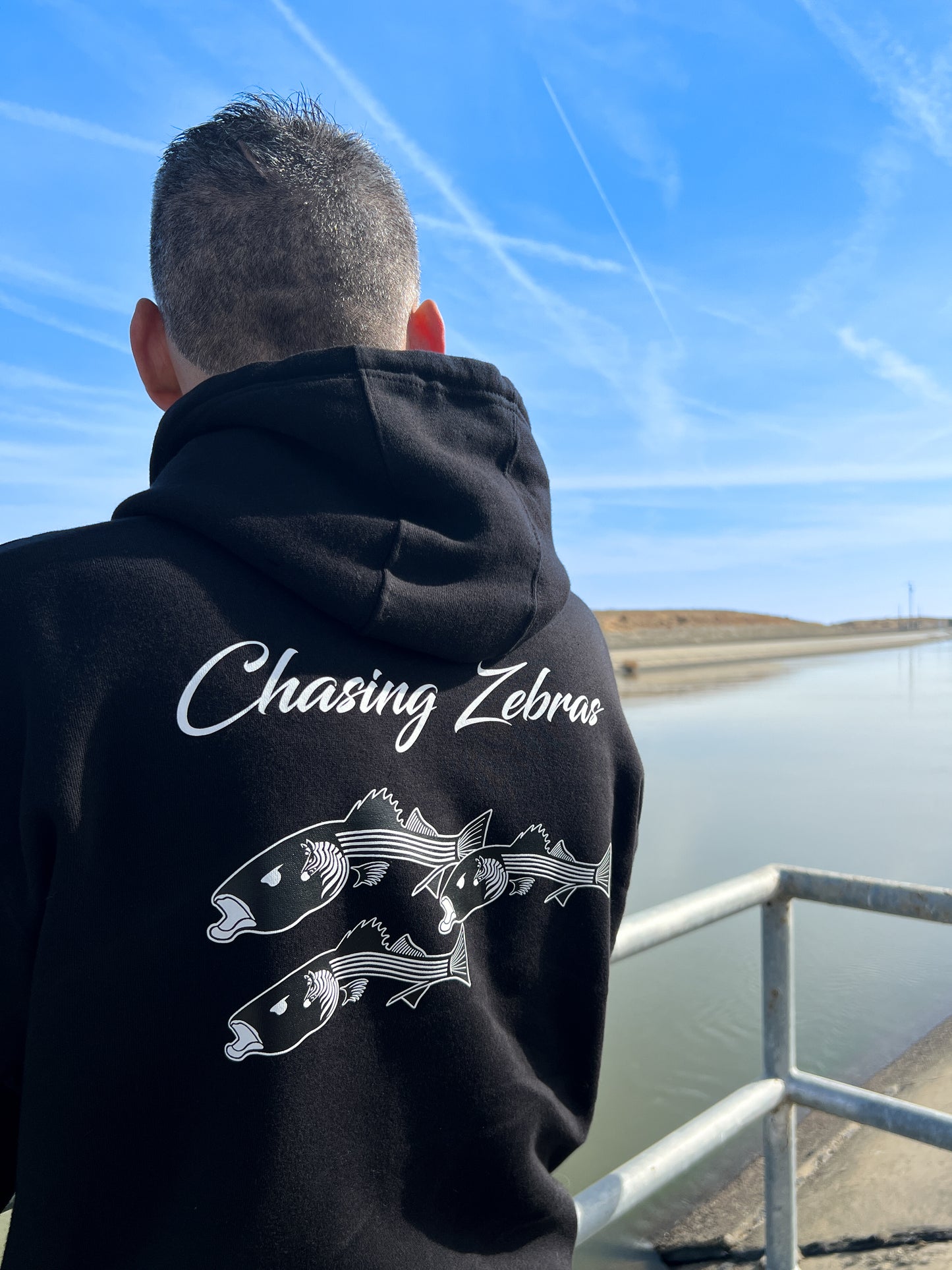 Person standing next to the California aqueduct with a black hoodie. On the back it says Chasing Zebras across the back and under it has a design of 3 striped bass in black and white and the striped bass have zebras on the side of their head