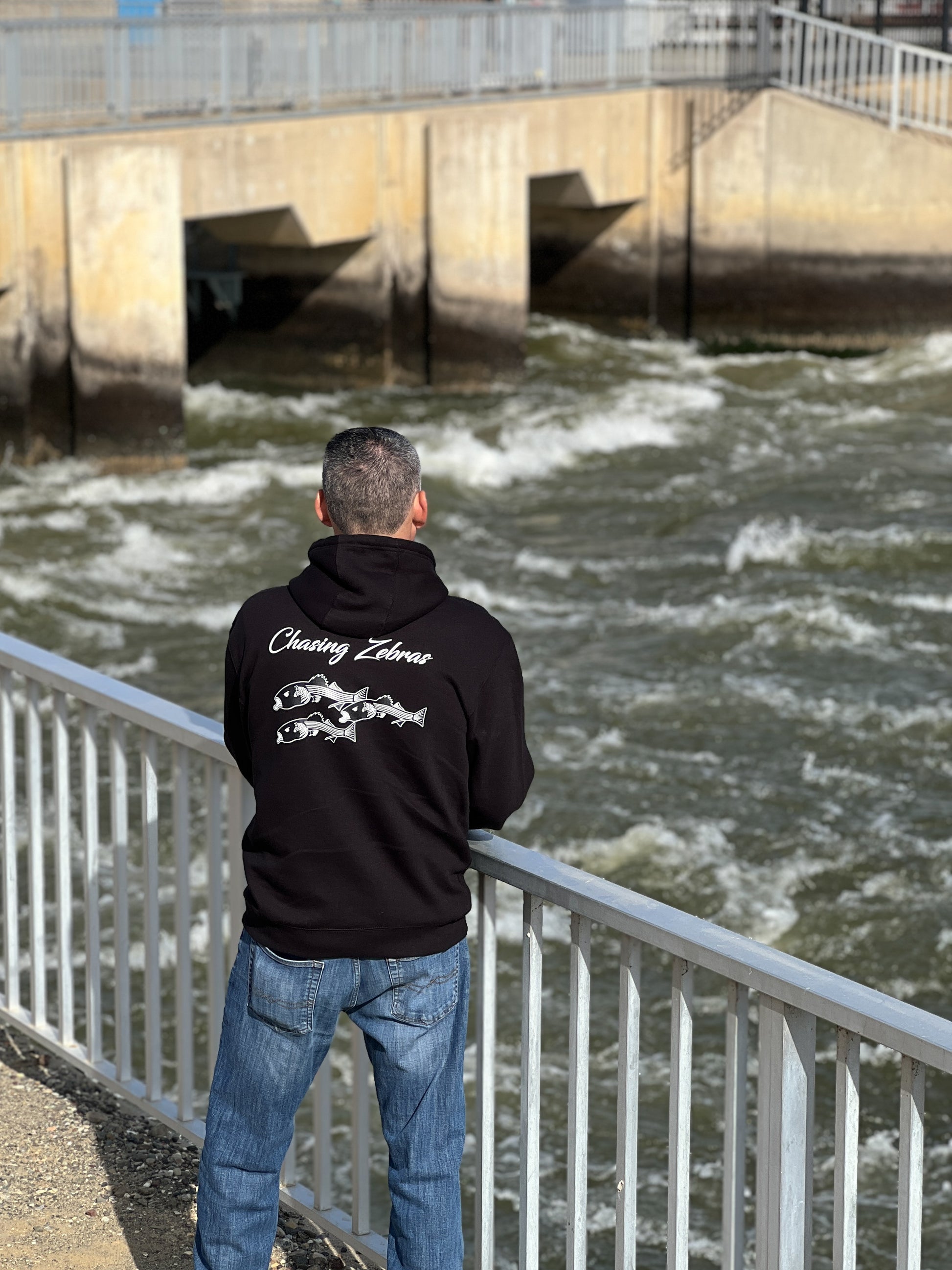 Person standing next to the California aqueduct with a black hoodie. On the back it says Chasing Zebras across the back and under it has a design of 3 striped bass in black and white and the striped bass have zebras on the side of their head