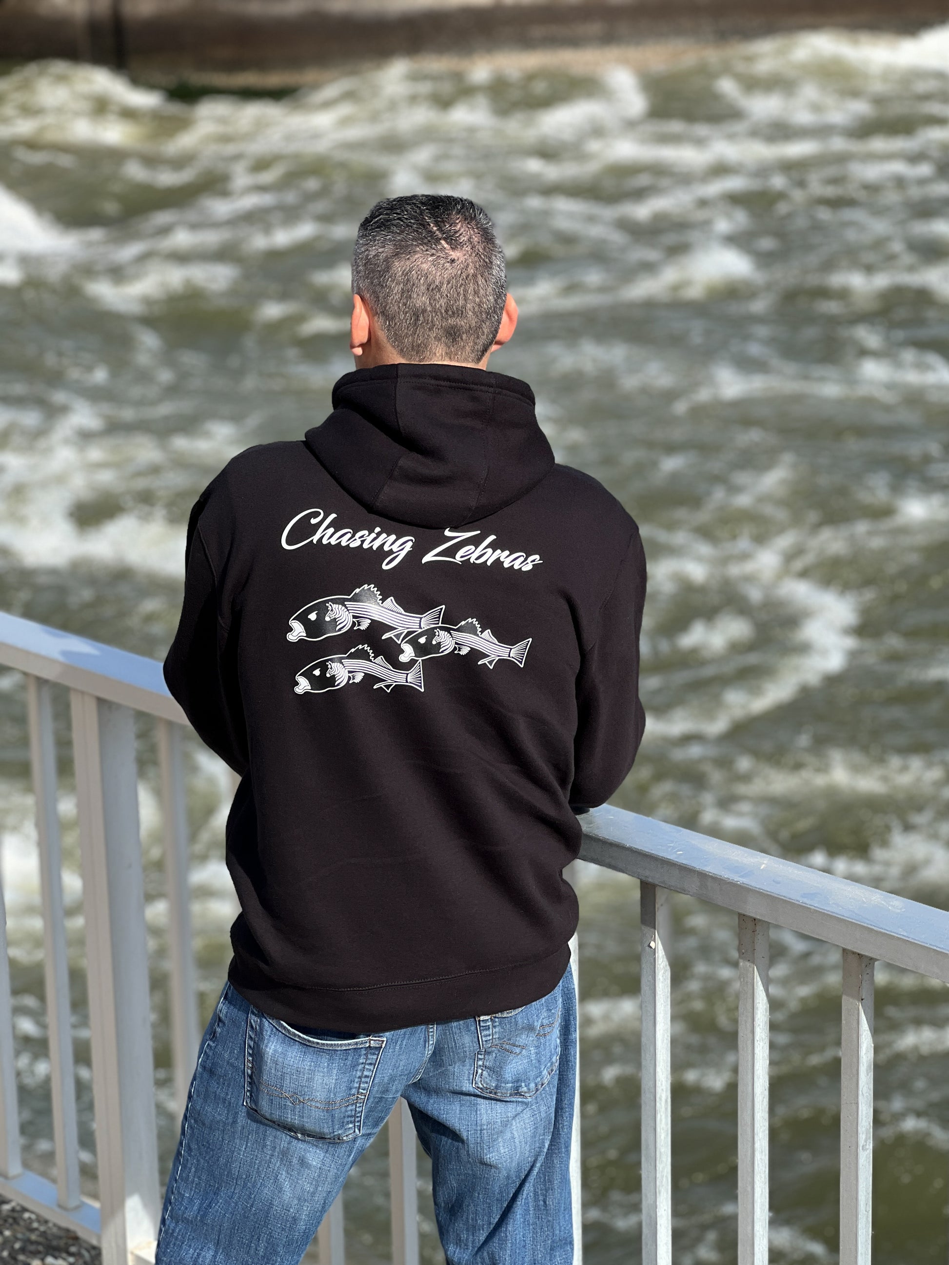 Person standing next to the California aqueduct with a black hoodie. On the back it says Chasing Zebras across the back and under it has a design of 3 striped bass in black and white and the striped bass have zebras on the side of their head