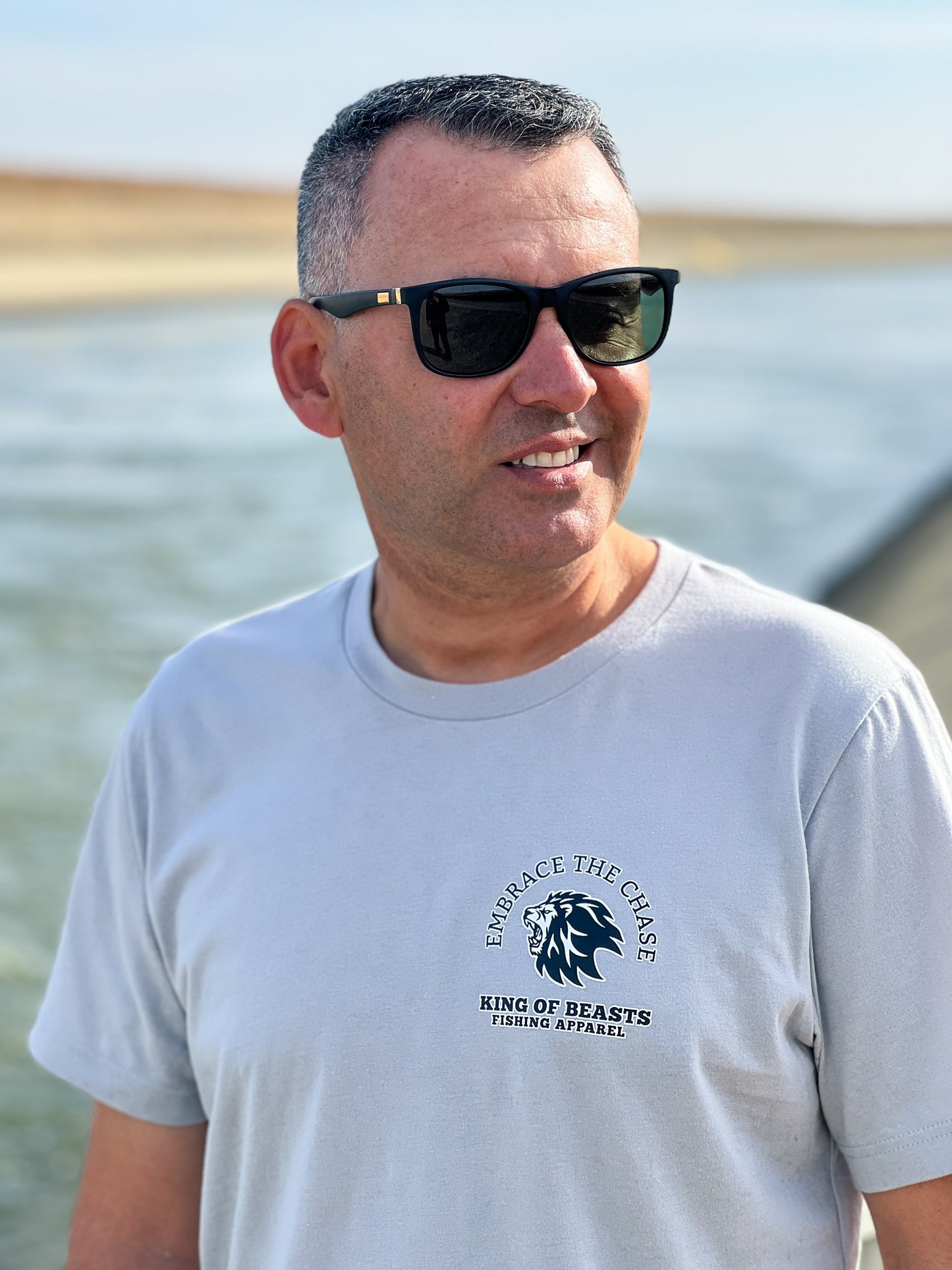 Person standing next to the California aqueduct with a light gray t shirt. On the left side of the chest it has a lion roaring and around the lion it says embrace the chase King Of Beasts Fishing Apparel.