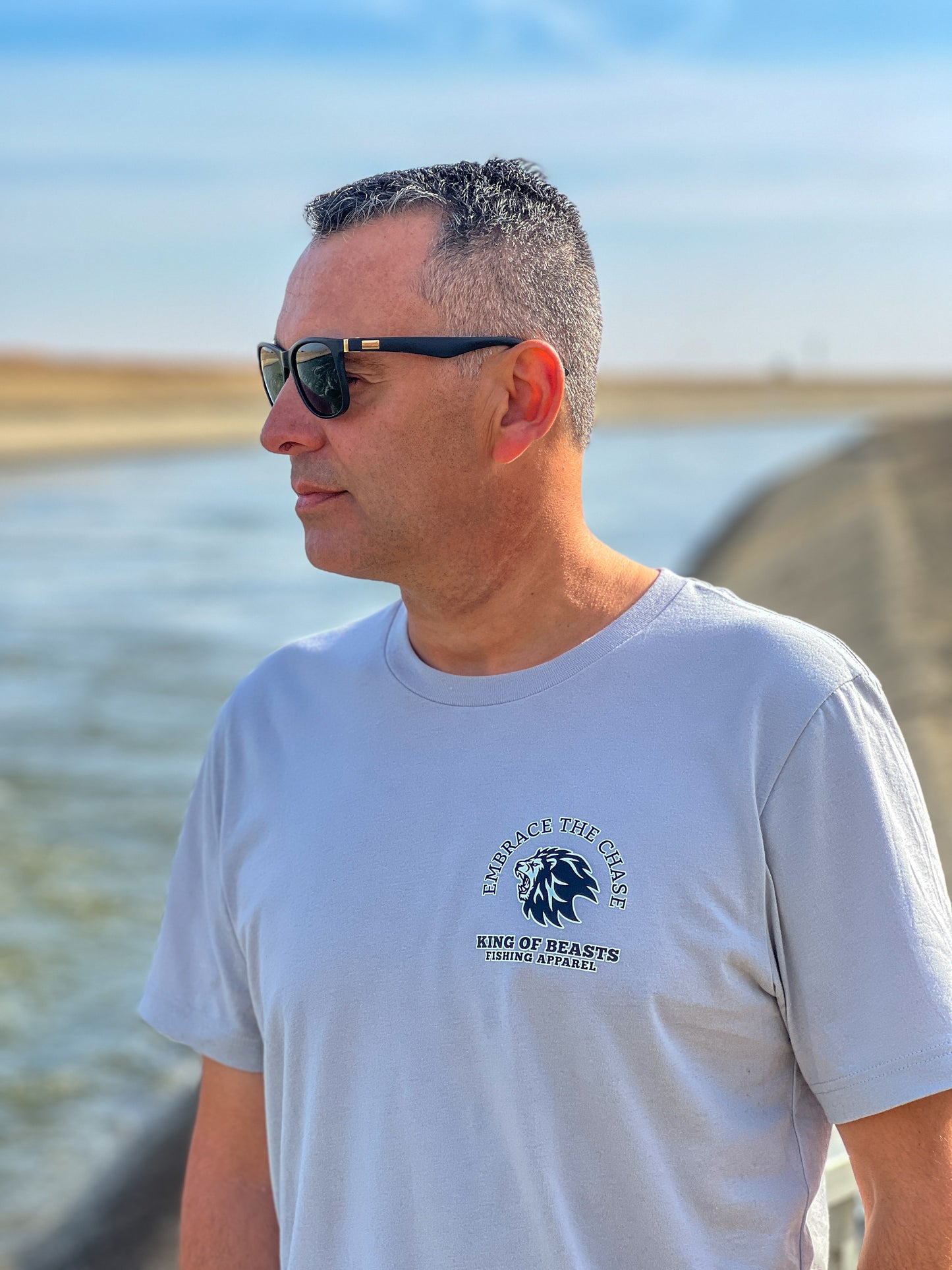 Person standing next to the California aqueduct with a light gray t shirt. On the left side of the chest it has a lion roaring and around the lion it says embrace the chase King Of Beasts Fishing Apparel.