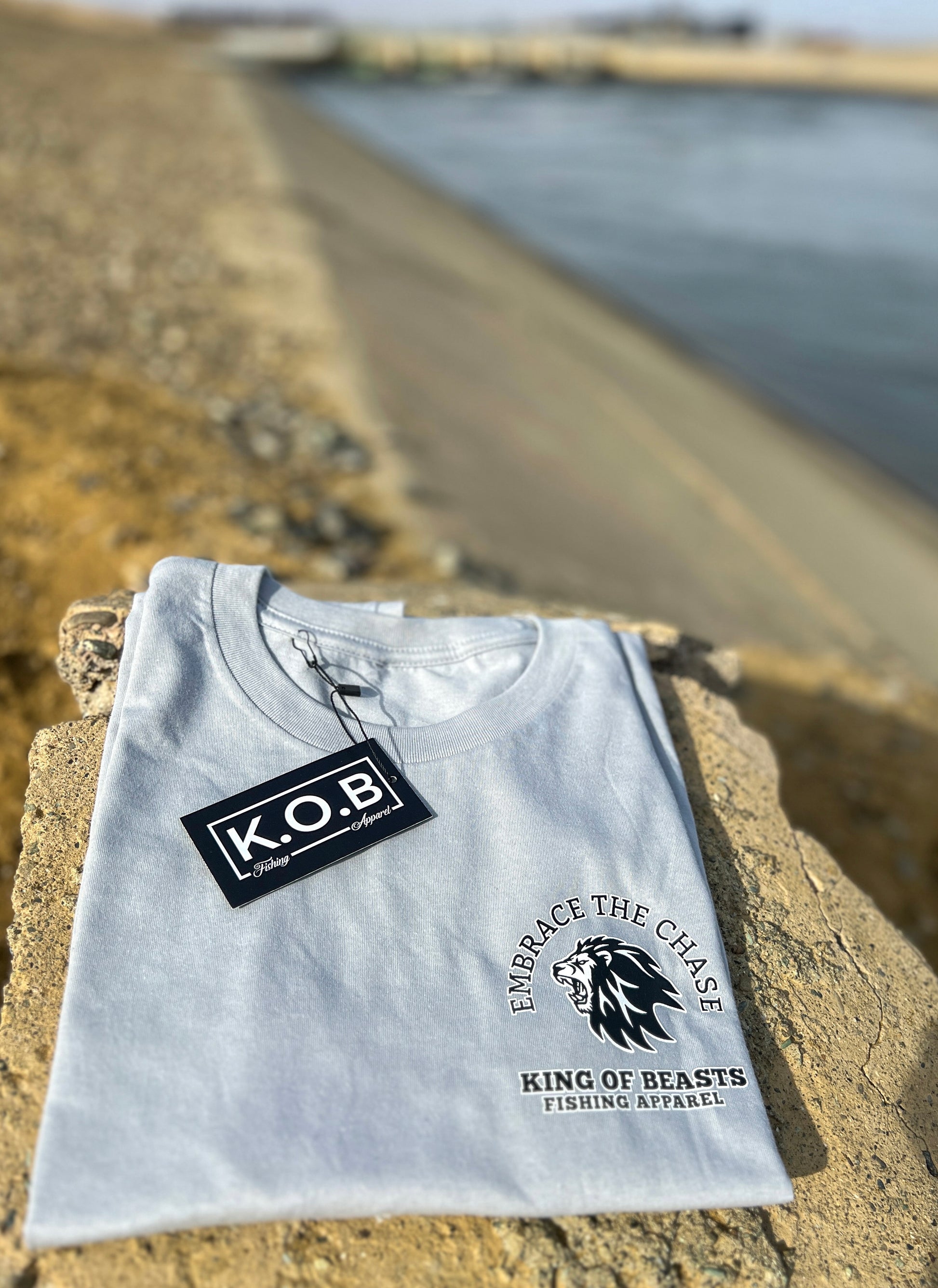 Light gray t shirt folded next to the California aqueduct. On the left side of the chest it has a lion roaring and around the lion it says embrace the chase King Of Beasts Fishing Apparel. It has a hang tag that says K.O.B Fishing Apparel