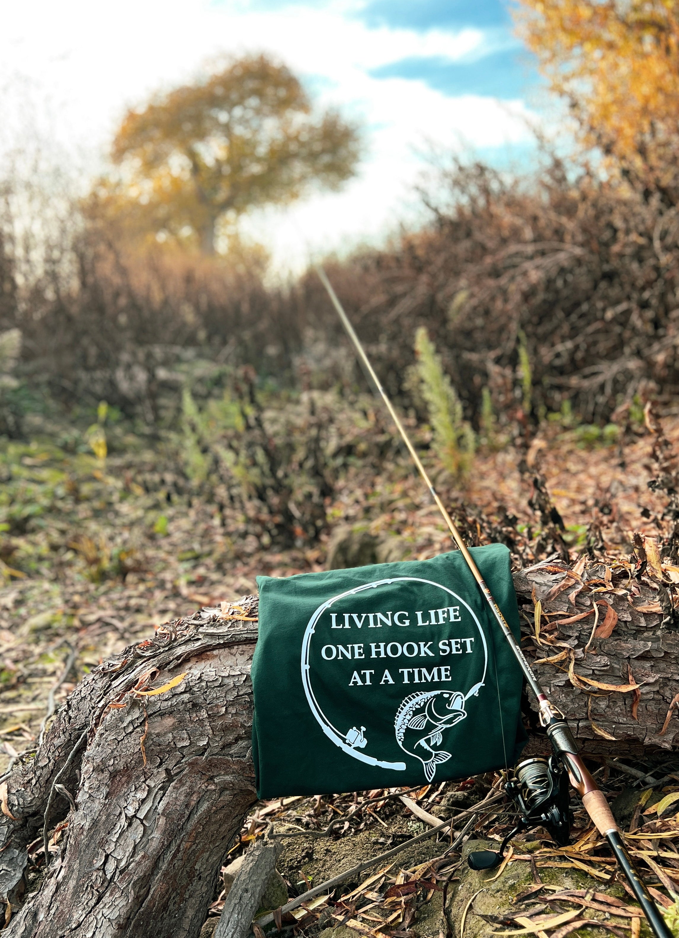 Forest green shirt on a tree branch with a fishing pole next to it. The design on the shirt is of a fishing rod bent in a circle and a large mouth bass at the end of it. In the middle of the design it says Living life one hook set at a time in capital letters