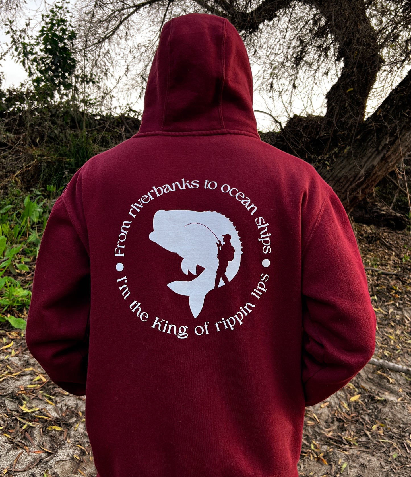 Person facing away with a burgundy hoodie. On the back of the hoodie in a circle it says From riverbanks to ocean ships I'm the king of rippin lips. in the middle of the words it has a large mouth bass and a silhouette of a man with a fishing pole and backpack on.