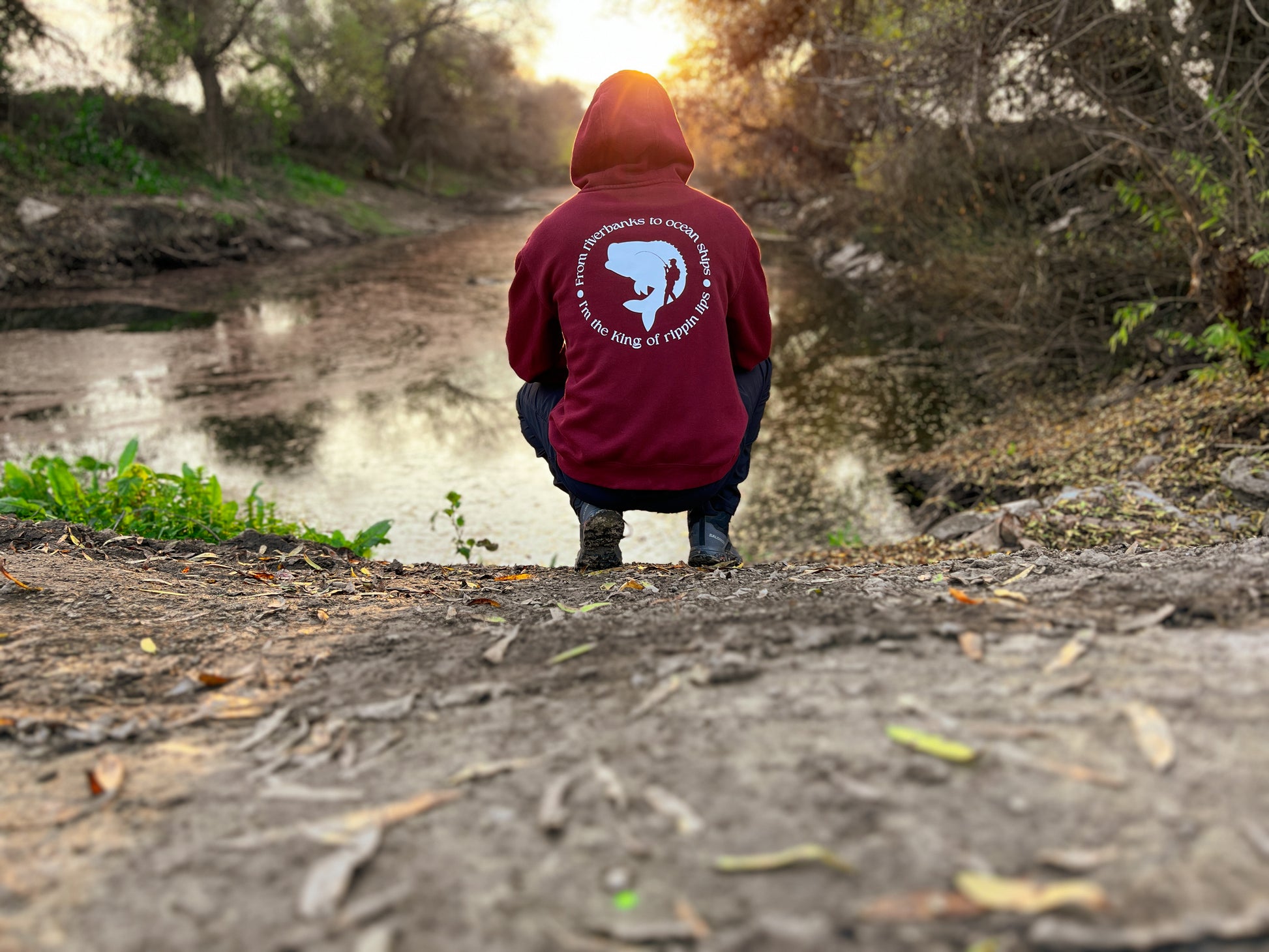 Person facing away with a burgundy hoodie. On the back of the hoodie in a circle it says From riverbanks to ocean ships I'm the king of rippin lips. in the middle of the words it has a large mouth bass and a silhouette of a man with a fishing pole and backpack on.