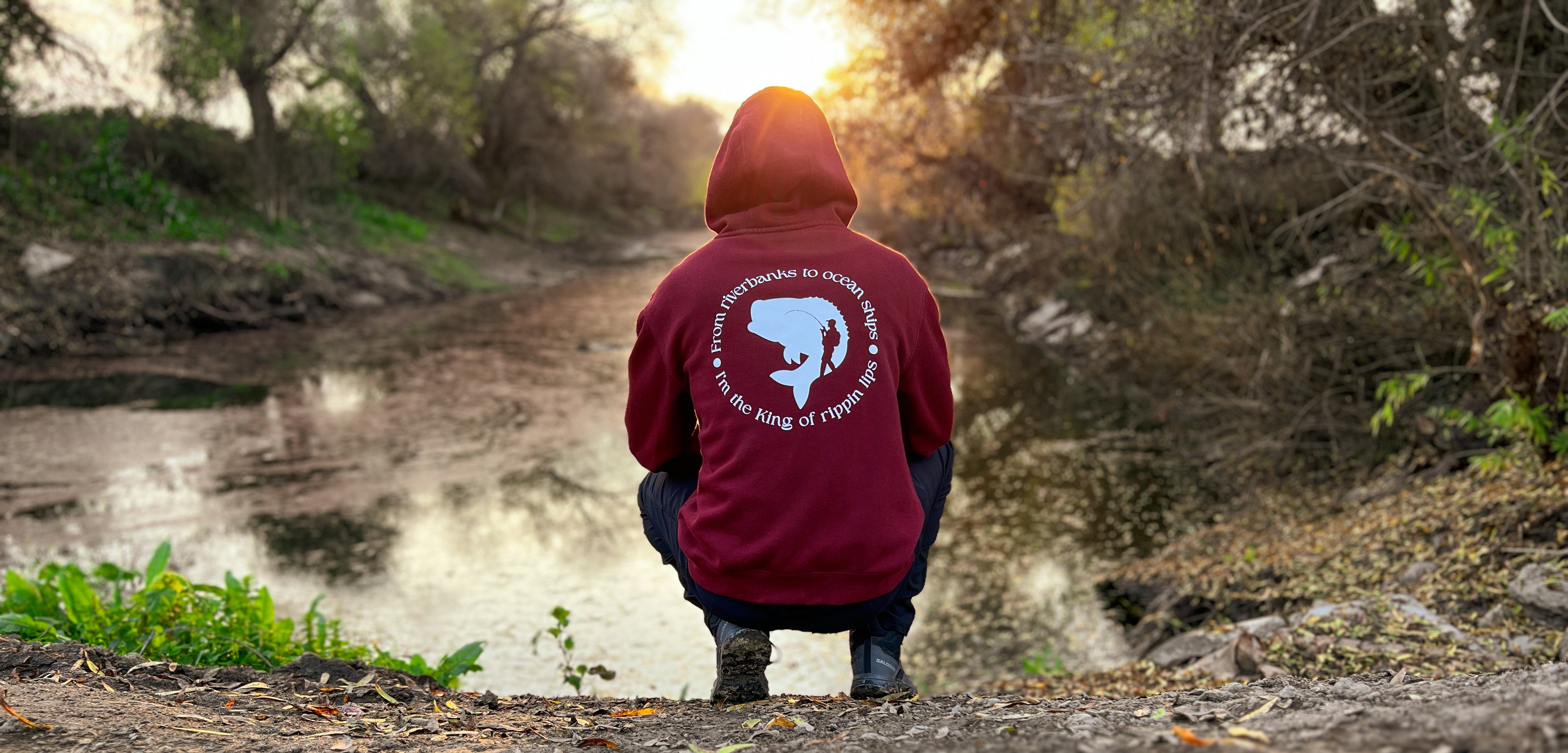 Person squatting down by a river looking into the sunset. He has on a burgundy hoodie that says from riverbanks to ocean ships I'm the king of rippin lips. 