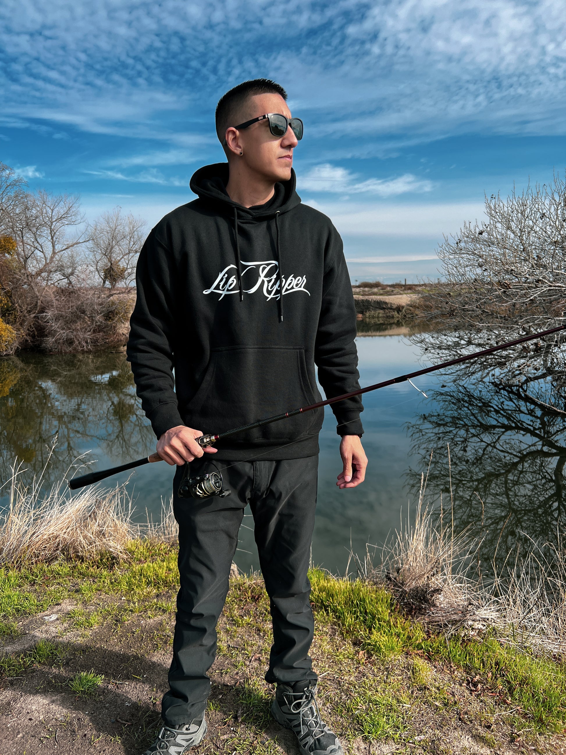 Person with sun glasses on by a river. He's holding a fishing pole with a black hoodie that says Lip Ripper