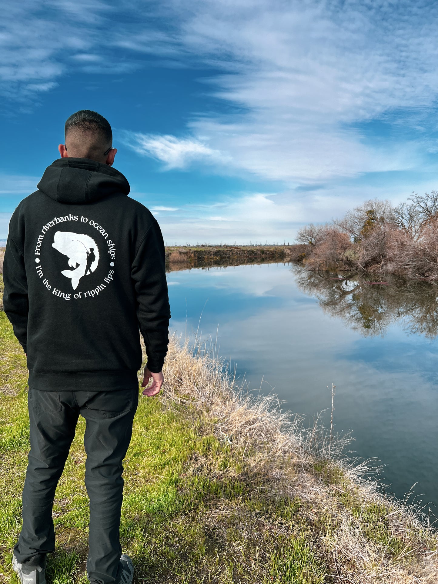 Person facing away with a black hoodie. On the back of the hoodie in a circle it says From riverbanks to ocean ships I'm the king of rippin lips. in the middle of the words it has a large mouth bass and a silhouette of a man with a fishing pole and backpack on.